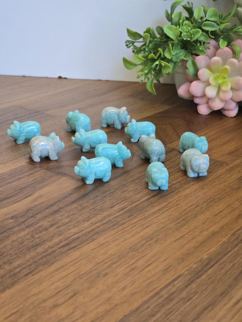 May include: A collection of twelve small, hand-carved animal figurines made from light blue, opaque stone. Each figurine is shaped like a pig with distinct ears, a snout, and four legs, arranged in a scattered formation across the surface.