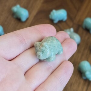 May include: A small, carved pig figurine made of polished, light green stone with subtle white marbling throughout. The sculpture features a rounded body, pointed ears, and a snout, showcasing a smooth, glossy texture and natural mineral variations.