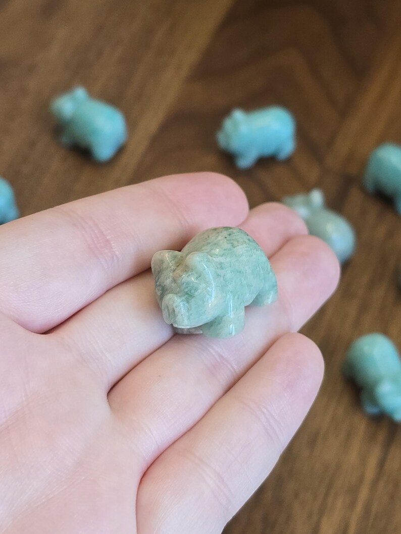 May include: A small, carved pig figurine made of polished, light green stone with subtle white marbling throughout. The sculpture features a rounded body, pointed ears, and a snout, showcasing a smooth, glossy texture and natural mineral variations.