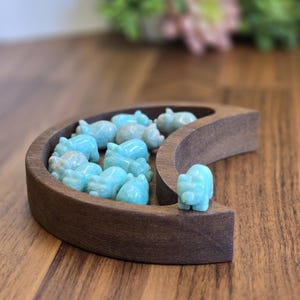 May include: A crescent moon-shaped wooden tray holding several small, carved light blue stone animal figurines. The tray is made of dark brown wood with a smooth finish, and the figurines are scattered inside the curved basin, with one placed on the outer edge.