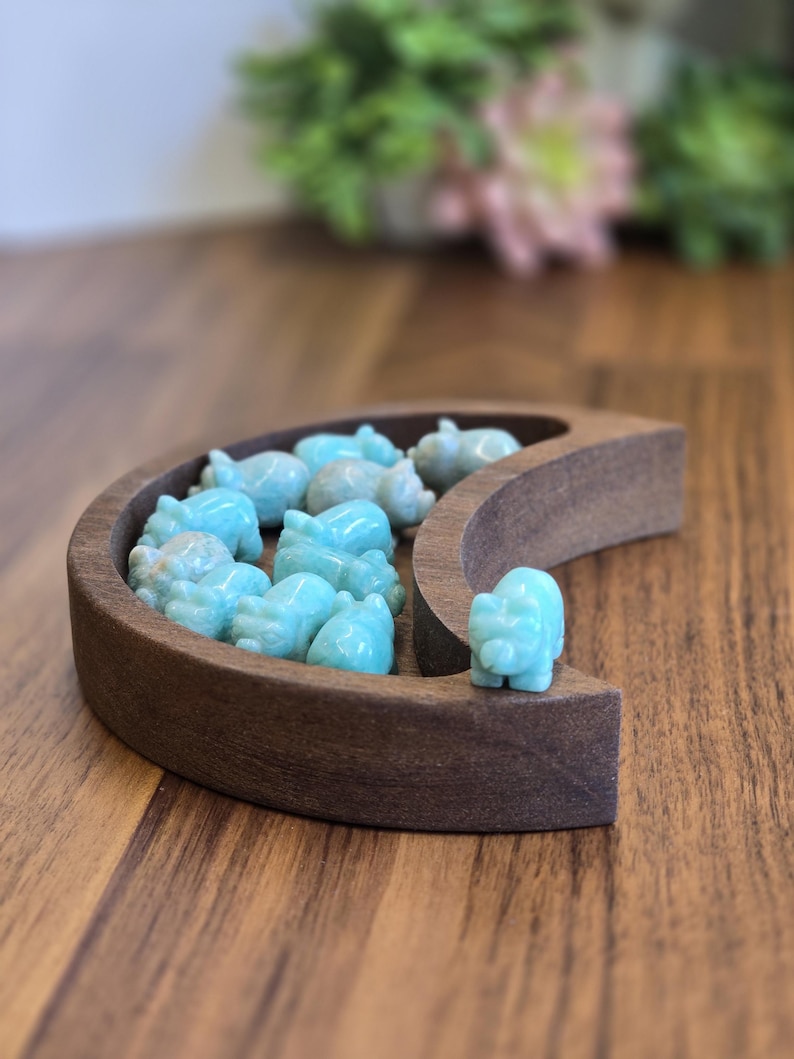 May include: A crescent moon-shaped wooden tray holding several small, carved light blue stone animal figurines. The tray is made of dark brown wood with a smooth finish, and the figurines are scattered inside the curved basin, with one placed on the outer edge.