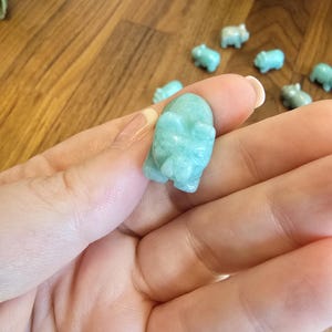 May include: A small, hand-carved figurine of a pig made from smooth, polished light blue-green stone. The carving features defined ears, a snout, and a rounded body, showcasing the natural color variations and texture of the mineral material.