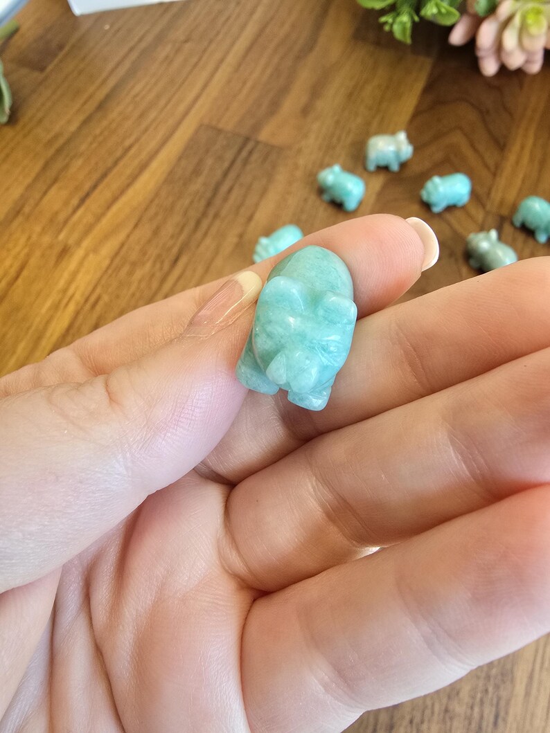 May include: A small, hand-carved figurine of a pig made from smooth, polished light blue-green stone. The carving features defined ears, a snout, and a rounded body, showcasing the natural color variations and texture of the mineral material.