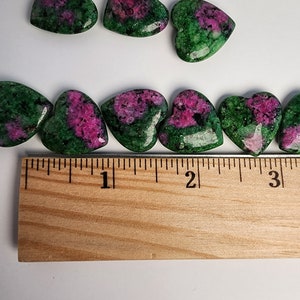 May include: A collection of green and pink heart-shaped gemstones, measuring approximately 1 inch in length, arranged on a wooden ruler.