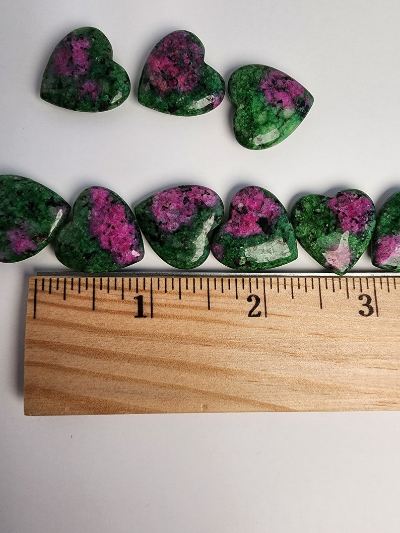 May include: A collection of green and pink heart-shaped gemstones, measuring approximately 1 inch in length, arranged on a wooden ruler.