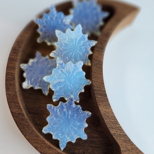 May include: A collection of six iridescent blue snowflake-shaped ornaments arranged in a crescent-shaped wooden bowl.