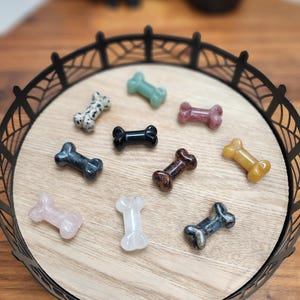 May include: A collection of ten polished crystal dog bone shapes in various colors and patterns, including black, white, green, red, brown, and yellow, displayed on a black metal tray with a spiderweb design.