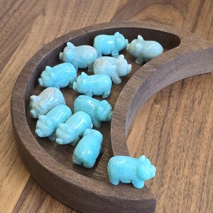 May include: A collection of eleven small, carved light blue stone pig figurines arranged inside a dark brown wooden crescent moon-shaped tray. Each pig is polished with a smooth finish, showing subtle variations in the natural stone color.