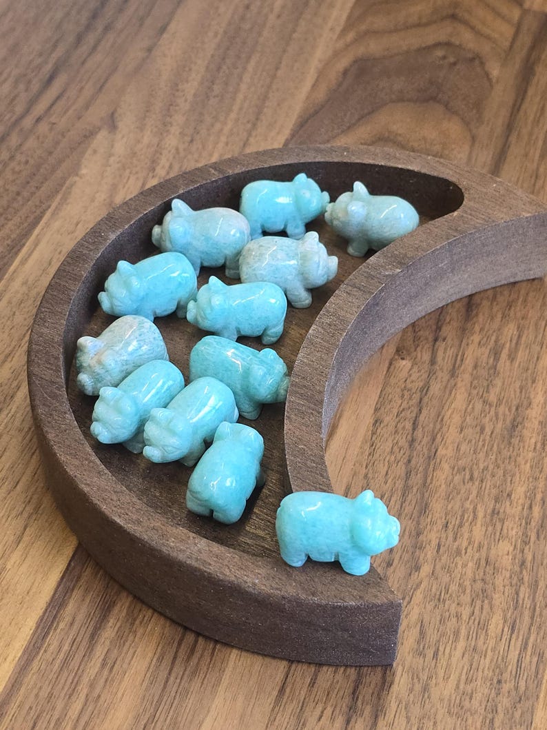 May include: A collection of eleven small, carved light blue stone pig figurines arranged inside a dark brown wooden crescent moon-shaped tray. Each pig is polished with a smooth finish, showing subtle variations in the natural stone color.