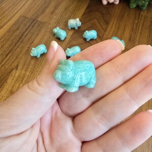 May include: A collection of small, carved light blue-green stone figurines shaped like pigs. One pig figurine is held in the foreground, while several other identical miniature pig carvings are scattered in the background.