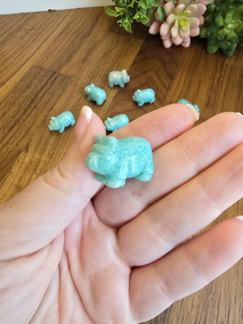 May include: A collection of small, carved light blue-green stone figurines shaped like pigs. One pig figurine is held in the foreground, while several other identical miniature pig carvings are scattered in the background.
