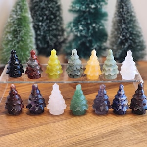 May include: A collection of 14 small, hand-carved crystal Christmas tree ornaments. The trees are made of various types of crystals, including amethyst, clear quartz, green aventurine, and red jasper. The trees are arranged in a row on a wooden surface.