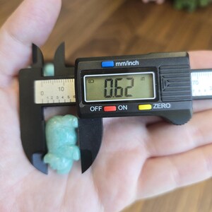 May include: A small, light green carved stone figurine shaped like an animal, measured by a black digital caliper showing a reading of 0.62 inches. The figurine has a smooth, polished surface and is held in place by the jaws of the measuring tool.