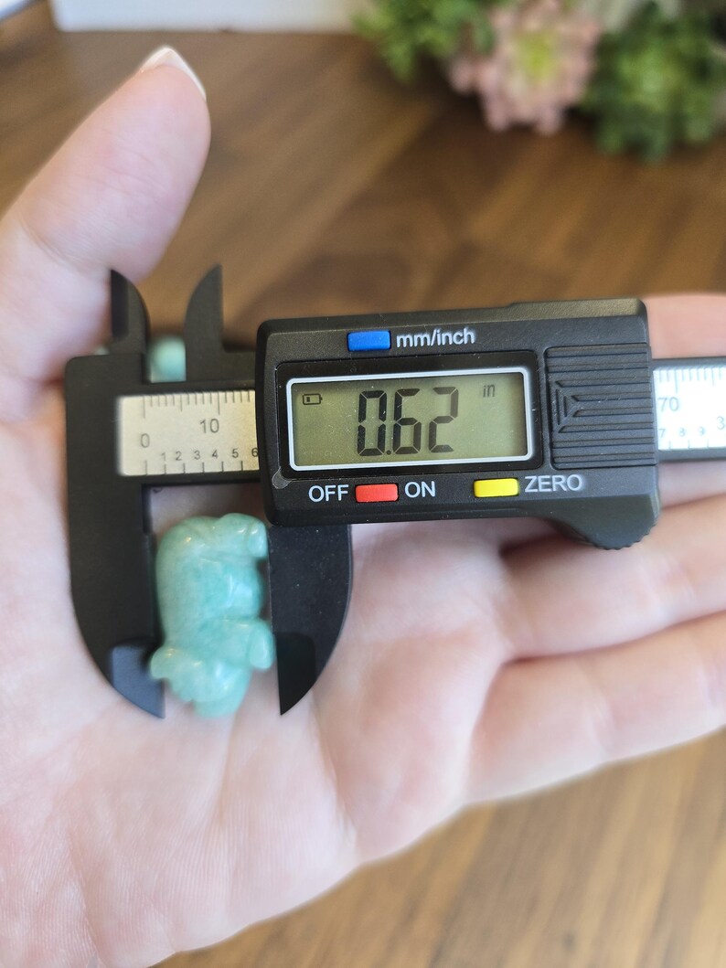 May include: A small, light green carved stone figurine shaped like an animal, measured by a black digital caliper showing a reading of 0.62 inches. The figurine has a smooth, polished surface and is held in place by the jaws of the measuring tool.