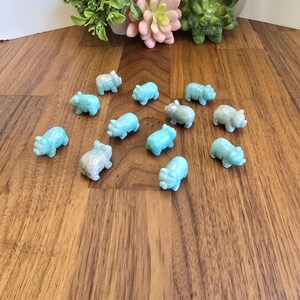 May include: A collection of twelve small, light blue, carved stone pig figurines arranged in a scattered formation. Each miniature animal sculpture features a smooth, polished surface with subtle variations in color and texture, depicting a standing pig.
