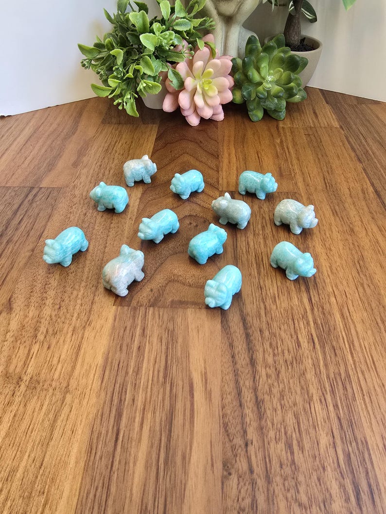 May include: A collection of twelve small, light blue, carved stone pig figurines arranged in a scattered formation. Each miniature animal sculpture features a smooth, polished surface with subtle variations in color and texture, depicting a standing pig.