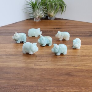 May include: A collection of seven small, hand-carved pig figurines made from smooth, pale mint-green stone. Each miniature animal features rounded ears, a snout, and a compact body, arranged in a scattered formation on a wooden surface.