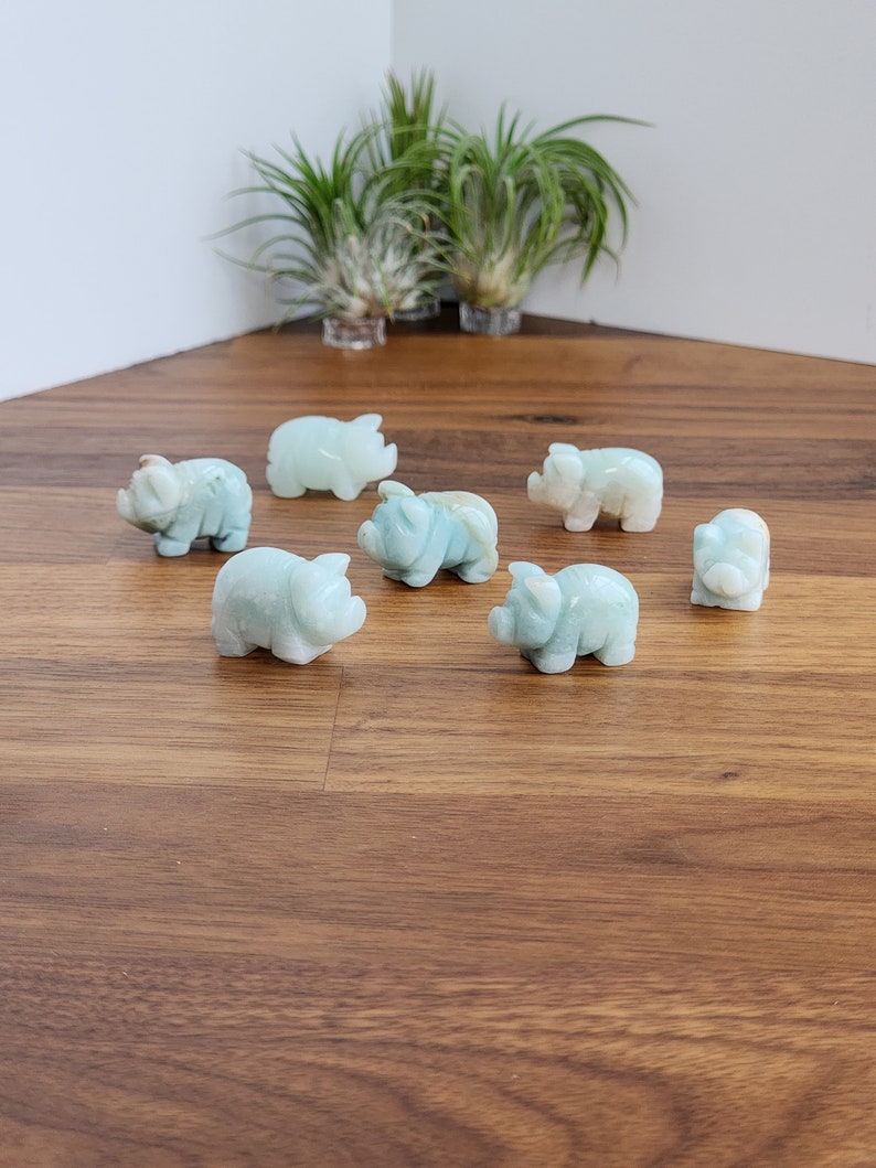 May include: A collection of seven small, hand-carved pig figurines made from smooth, pale mint-green stone. Each miniature animal features rounded ears, a snout, and a compact body, arranged in a scattered formation on a wooden surface.