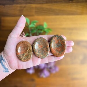 May include: Three oval-shaped unakite stones with a mix of orange, red, and green hues. The stones are smooth and polished, showcasing their natural patterns. The stones are held in a hand, with a wooden background.