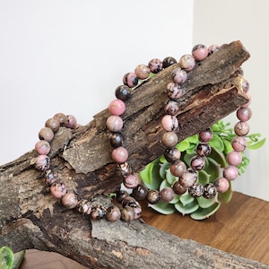 May include: Two beaded bracelets with pink, brown, and black stones. The bracelets are draped over a piece of weathered wood. The beads are round and appear to be made of a natural stone. The background is a neutral white and green.