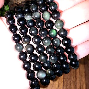 May include: A strand of polished, round, black obsidian beads. The beads have a glossy finish and subtle iridescent green and silver inclusions. The beads are strung together, forming a necklace or bracelet. The background is a wooden surface.