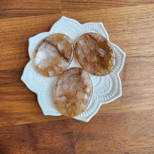 May include: Three oval-shaped, polished stones with brown and white inclusions rest on a white, flower-shaped dish. The stones have a translucent quality, and the dish is placed on a wooden surface.