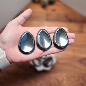 May include: Three polished, teardrop-shaped, hematite stones are held in a hand. The stones have a dark, metallic gray color and reflect the surrounding environment. The background is a wooden surface.