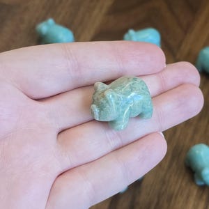 May include: A small, polished, light green stone figurine carved into the shape of a pig. The surface of the stone has a smooth, glossy texture with subtle variations in color and natural mineral patterns throughout the body of the animal.