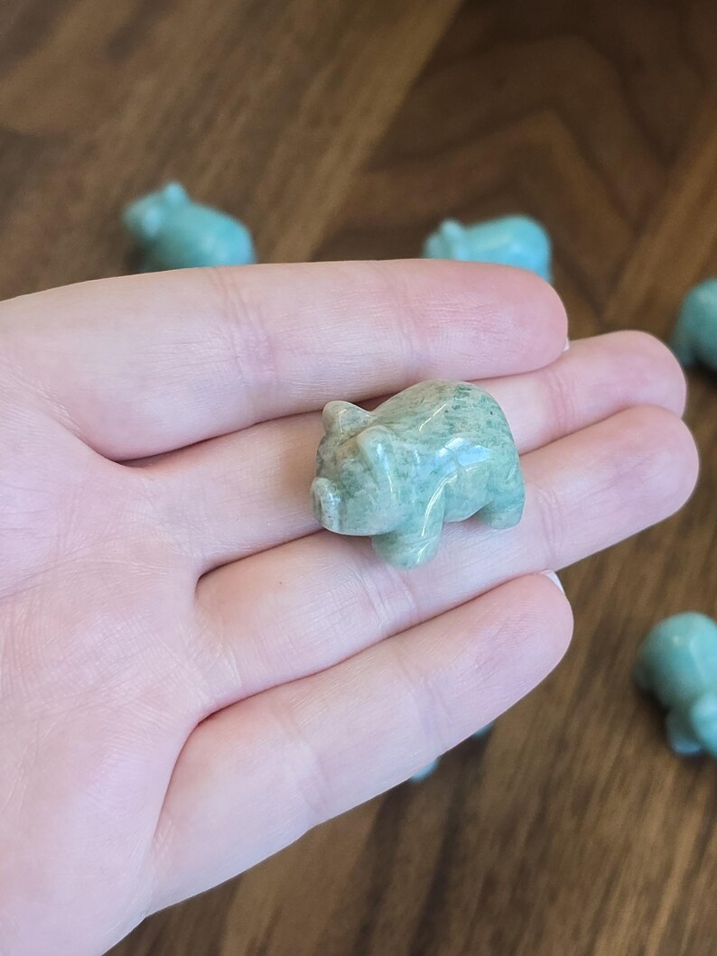 May include: A small, polished, light green stone figurine carved into the shape of a pig. The surface of the stone has a smooth, glossy texture with subtle variations in color and natural mineral patterns throughout the body of the animal.