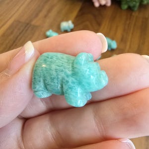 May include: A small, hand-carved figurine of a pig made from polished amazonite stone. The stone features a mottled, opaque teal and light green color palette with natural white veining throughout the smooth, rounded surface of the animal sculpture.
