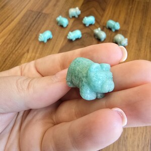 May include: A small, hand-carved figurine of a pig made from light green, semi-translucent stone. The pig has rounded features, visible ears, and a snout. Several identical miniature pig carvings are scattered in the background on a wooden surface.