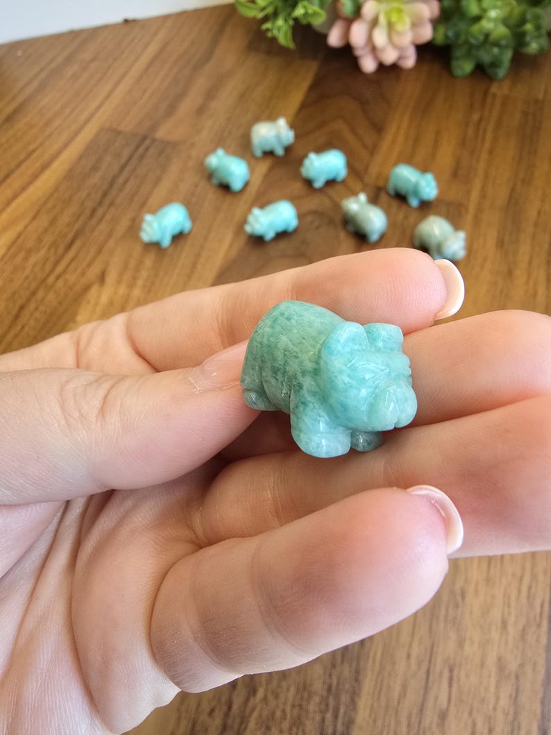 May include: A small, hand-carved figurine of a pig made from light green, semi-translucent stone. The pig has rounded features, visible ears, and a snout. Several identical miniature pig carvings are scattered in the background on a wooden surface.