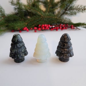May include: Three carved gemstone Christmas tree figurines in shades of green, white, and black. The trees are arranged in a row against a white background, with a sprig of evergreen and red berries in the background. The trees are a festive holiday decoration.
