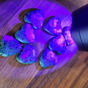 May include: A group of eight heart-shaped stones, illuminated by a UV light, showing a vibrant mix of green, purple, and pink hues.