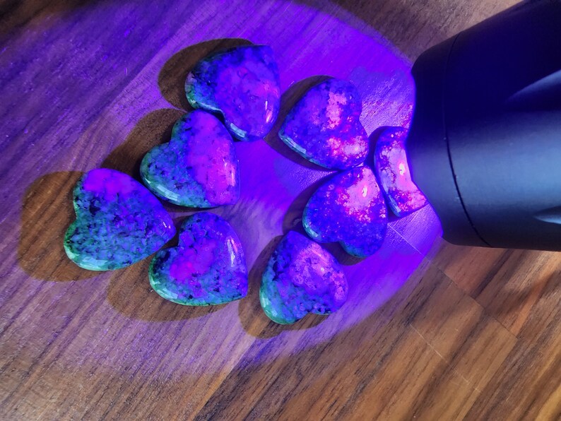 May include: A group of eight heart-shaped stones, illuminated by a UV light, showing a vibrant mix of green, purple, and pink hues.