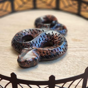 May include: A brown and copper-colored snake figurine with carved details, coiled on a wooden surface. The snake is the primary focus, with a decorative metal tray in the background.