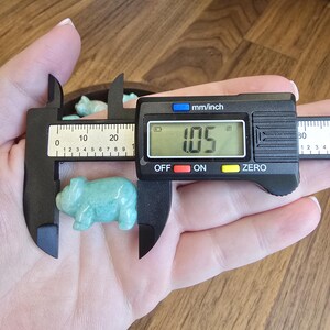 May include: A small, light green, carved stone pig figurine being measured by a black digital caliper. The digital display on the caliper clearly shows a measurement of 1.05 inches, indicating the length of the smooth, polished stone animal.