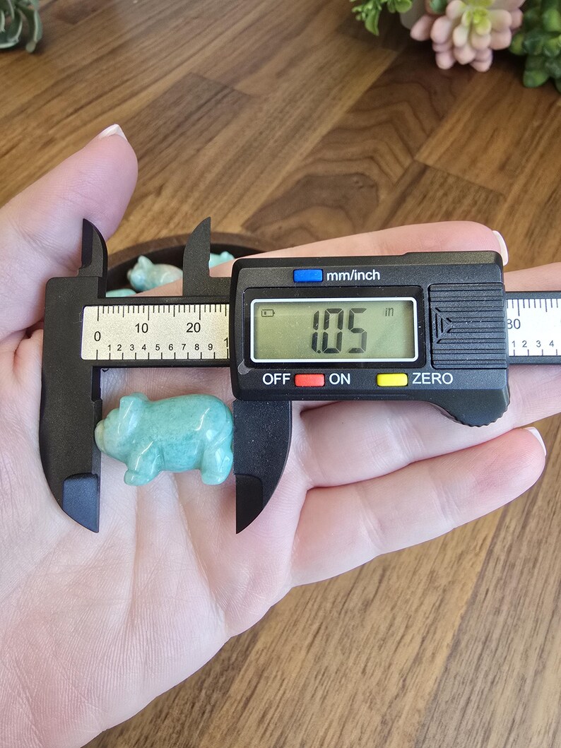 May include: A small, light green, carved stone pig figurine being measured by a black digital caliper. The digital display on the caliper clearly shows a measurement of 1.05 inches, indicating the length of the smooth, polished stone animal.