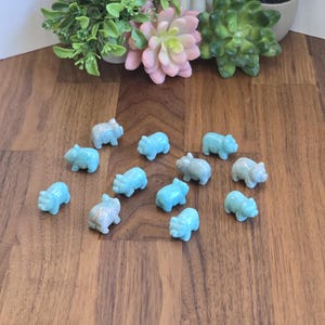 May include: A collection of twelve small, carved light blue stone figurines shaped like pigs. Each miniature animal figure features a smooth, polished surface with subtle variations in color and texture, arranged in a scattered formation.