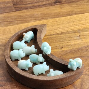 May include: A crescent moon-shaped wooden tray containing nine small, carved light green stone pig figurines. The pigs are arranged within the curved basin of the dark brown wooden tray, each displaying smooth surfaces and subtle variations in color.