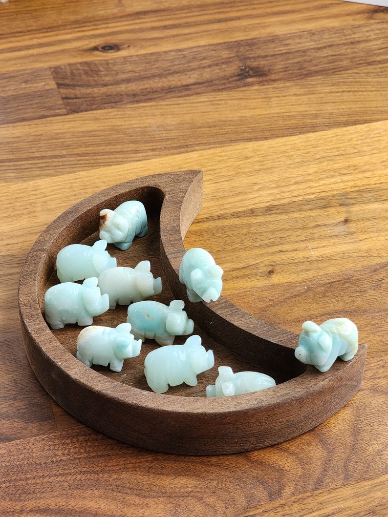 May include: A crescent moon-shaped wooden tray containing nine small, carved light green stone pig figurines. The pigs are arranged within the curved basin of the dark brown wooden tray, each displaying smooth surfaces and subtle variations in color.