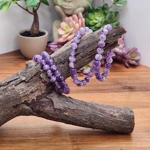 May include: Three amethyst gemstone bracelets stacked on a piece of driftwood. The bracelets are made of small, round beads.