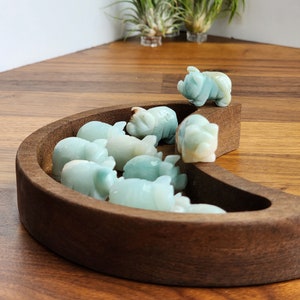 May include: A collection of small, light blue-green carved stone pig figurines arranged inside a crescent-shaped dark brown wooden bowl. The smooth, polished stone animals are clustered together, with one pig perched on the edge of the wooden rim.