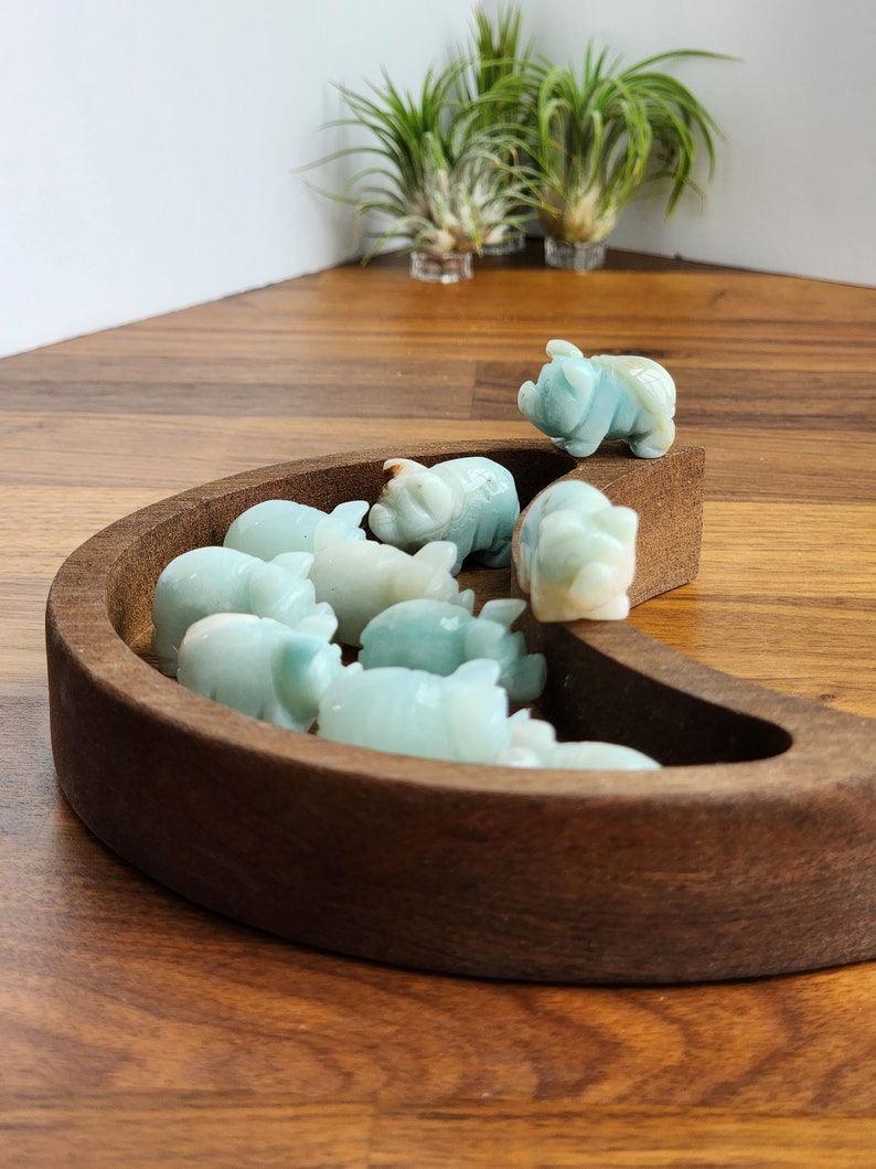 May include: A collection of small, light blue-green carved stone pig figurines arranged inside a crescent-shaped dark brown wooden bowl. The smooth, polished stone animals are clustered together, with one pig perched on the edge of the wooden rim.