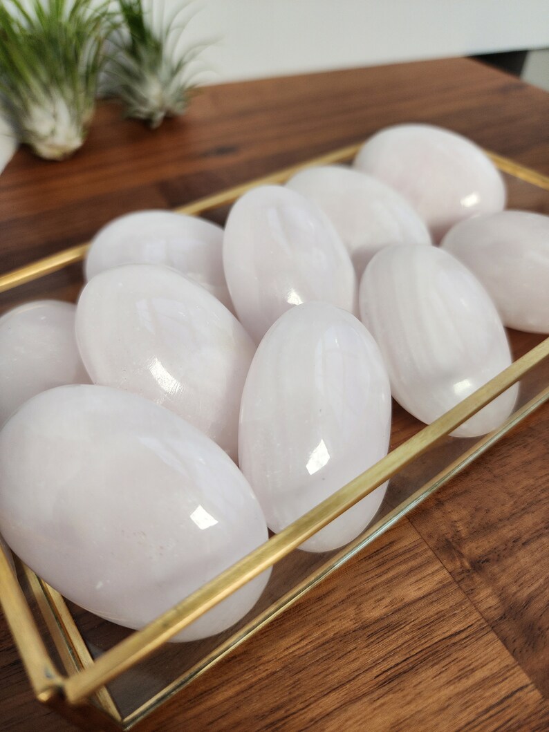 K&ouml;nnte beinhalten: Eine Sammlung glatter, wei&szlig;er Selenitsteine in einer Glas- und Goldschale. Die Steine sind oval geformt und haben einen dezenten Schimmer.