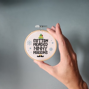May include: A white embroidery hoop with a cross-stitch design featuring a green elf hat, snowflakes, and the text "Cotton Headed Ninny Muggins".