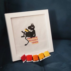 Könnte beinhalten: Ein weiß gerahmter Kreuzstich mit einer schwarzen Silhouette eines Dämons, der eine Banane hält. Der Text "Emotional Support Demon" ist in Rot und Orange unter dem Dämon gestickt.