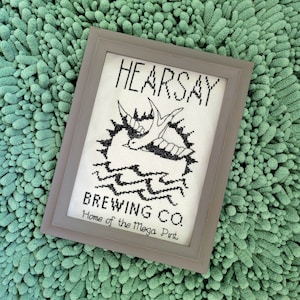 May include: A framed cross-stitch design featuring the text "HEARSAY BREWING CO. Home of the Mega Pint" with a stylized bird and waves.