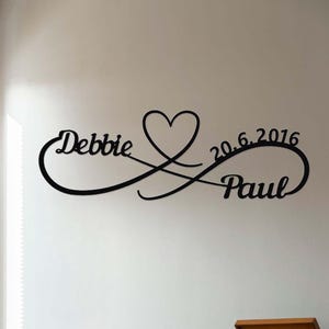 以下が含まれることがあります： 黒い金属製の壁掛けアート。名前「Debbie」と「Paul」がハートと無限大の記号で絡み合っています。「20.6.2016」の日付も含まれています。結婚式や記念日のギフトに最適な装飾品です。