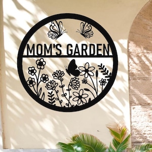 Könnte beinhalten: Schwarzes rundes Schild aus Metall mit dem Text "MOM'S GARDEN" in einem Banner oben. Die untere Hälfte des Kreises zeigt eine Silhouette von Blumen und Schmetterlingen.
